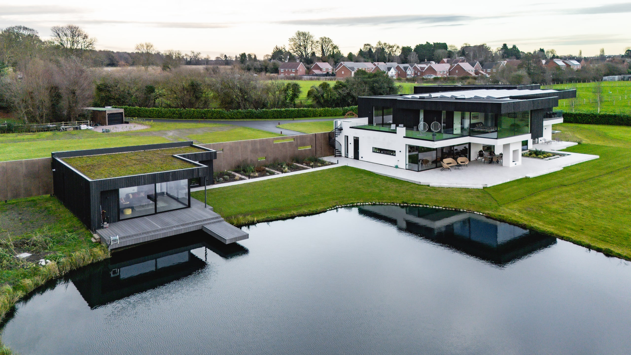 Modern two-story white and black residence with separate lakeside pavilion featuring green roof, set in rural landscape
